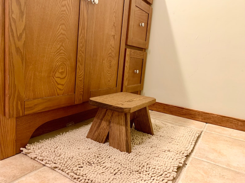 May include: A wooden step stool with a natural brown finish. The stool is sitting on a white shag rug in a bathroom.