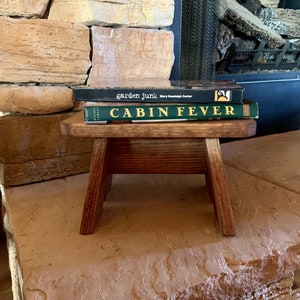 May include: A small wooden stool with two books stacked on top. The top book is titled "Cabin Fever" and the bottom book is titled "garden junk" by Mary Randolph Carter.