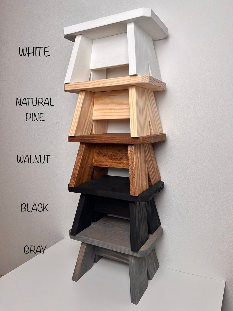 May include: Five wooden step stools stacked on top of each other. The stools are painted in different colors: white, natural pine, walnut, black, and gray.