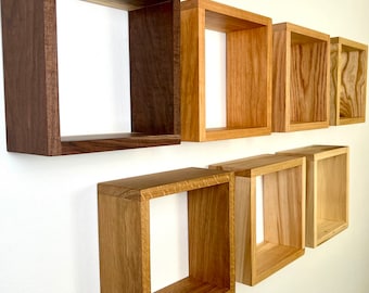 May include: A set of eight square wooden shelves in various shades of brown, mounted on a white wall. The shelves are made of different types of wood, including walnut and oak.