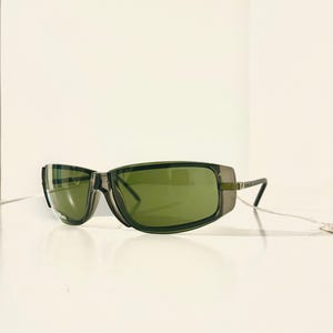 May include: Dark green sunglasses with a rectangular frame and green tinted lenses. The sunglasses have a sleek, modern design with a subtle metallic accent on the temples. The lenses are slightly curved, offering a stylish and functional accessory.