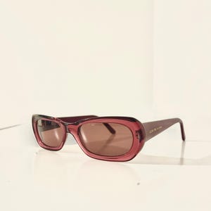 May include: Oval-shaped sunglasses in a deep burgundy hue. The frames have a translucent quality, and the lenses are a warm brown. The temples are slender and feature subtle text. A stylish accessory for eye protection.