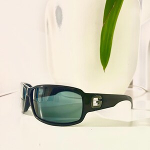 May include: Black sunglasses with a rectangular frame and tinted lenses. The sunglasses have a small silver emblem on the temples. The sunglasses are resting on a white surface.