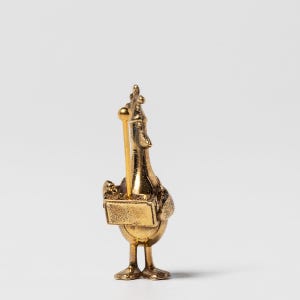 May include: A gold-coloured figurine of a duck wearing a crown and holding a sceptre. The duck is standing on a small platform.