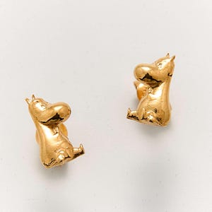 May include: A pair of gold-coloured hippo stud earrings. Each earring features a detailed, seated hippo design with a smooth, reflective surface. The earrings are set against a plain white background, highlighting their form and colour.