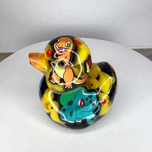 May include: A clear resin rubber duck containing multiple small yellow rubber ducks inside. The exterior surface features colorful graphic illustrations of an orange lizard-like creature and a teal frog-like creature with a large green bulb on its back.