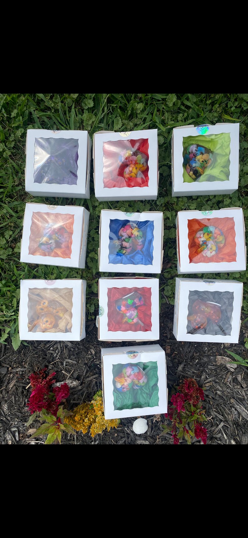 May include: Twelve small white boxes with clear plastic windows, each containing a colorful, confetti-like mixture. The boxes are arranged in three rows of four, on a bed of brown mulch.