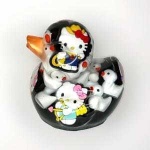 May include: A clear resin rubber duck containing multiple white rubber ducks and two Hello Kitty figures. One Hello Kitty wears a yellow hat and blue outfit, while the other has a pink bow and holds a yellow bow and arrow. The duck has an orange beak.