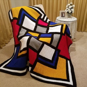 May include: A colorful crocheted throw blanket with a geometric pattern in shades of red, yellow, blue, white, and gray. The blanket is draped over a chair and has a black border.