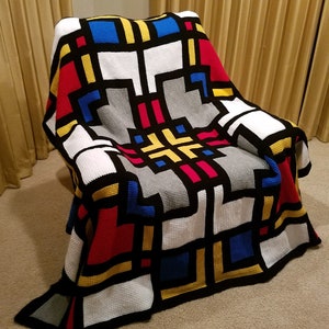 Könnte beinhalten: Eine gehäkelte Decke mit einem geometrischen Muster in Rot, Gelb, Blau, Schwarz, Weiß und Grau. Die Decke ist gefaltet und über einen Stuhl gelegt.