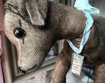 May include: A brown fabric dog figurine with a dark brown stitched nose and black button eyes. It wears a white lace collar featuring a small rectangular metal pendant with a portrait of a child and the German text Sie ähnelt dem Großvater.