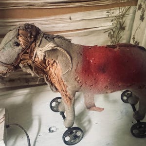 Könnte beinhalten: Ein antikes Spielzeugpferd mit abgenutztem Aussehen. Das Pferd ist überwiegend grau mit einem roten Bereich auf dem Rücken. Es hat ein Halfter und ist auf vier kleinen schwarzen Rädern montiert.