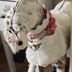 May include: A handcrafted fabric rocking horse figure featuring a distressed white and beige textured exterior. The horse wears a white collar with eyelet lace trim, a small embroidered floral detail, and a ruffled red and white checkered ribbon.