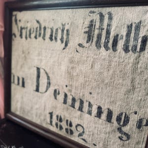 Könnte beinhalten: Ein gerahmtes Stück Stoff mit dem Text "Friedrich Merker in Deining 1882" in schwarzer Tinte gedruckt.