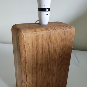 May include: A wooden table lamp featuring a rectangular base and a white bulb socket. The base is made of light brown wood with a natural grain pattern. The lamp is designed for indoor use and has a simple, modern aesthetic.