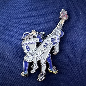 May include: A baseball pin featuring two players in action. One player is wearing the number 8 and the name "ERNANDEZ" on the back of his jersey. The other player is wearing the number 44 and the name "PAGES" on the back of his jersey. The pin has blue, white, and yellow accents.