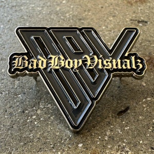 May include: A black and silver metal pin with the text "Bad Boy Visualz" in gold lettering. The pin is shaped like a triangle with three lines.