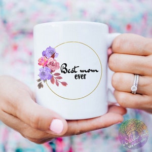 May include: White ceramic mug with a gold circle and floral design. The text "Best mom ever" is written in black inside the circle.