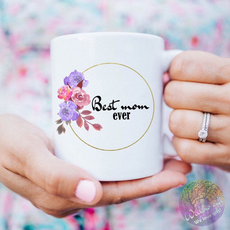 May include: White ceramic mug with a gold circle and floral design. The text "Best mom ever" is written in black inside the circle.