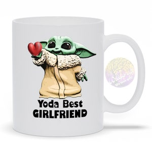 Puede incluir: Taza de cer&aacute;mica blanca con un personaje de dibujos animados verde y marr&oacute;n que sostiene un coraz&oacute;n rojo. El texto "Yoda Best Girlfriend" est&aacute; impreso en la taza.