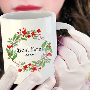 May include: White ceramic mug with a floral wreath design in shades of green, red, and blue. The wreath surrounds the text "Best Mom ever".