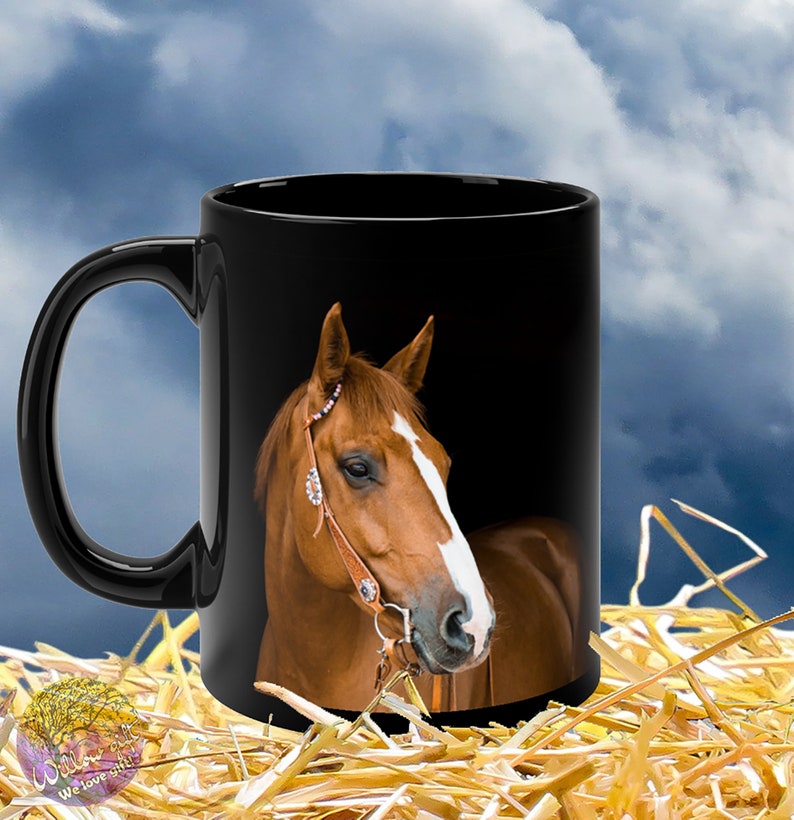 May include: Black coffee mug with a brown horse with a white stripe on its face. The horse is wearing a leather bridle with a silver buckle. The mug is sitting on a bed of straw.