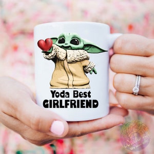 Puede incluir: Taza de cer&aacute;mica blanca con un personaje de dibujos animados verde que sostiene un coraz&oacute;n rojo. El texto "Yoda Best Girlfriend" est&aacute; impreso en negro en la taza.