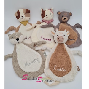 Könnte beinhalten: Eine Sammlung weicher, gestrickter Baby-Kuscheldecken in Tierform. Das Sortiment umfasst einen Hund, eine Katze, einen Bären, eine Kuh und einen Fuchs, jeweils mit gestickten Namen. Die Decken sind in neutralen Farben gehalten und haben strukturierte Details und angebrachte Arme.