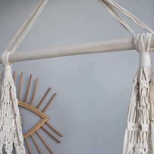Puede incluir: Una silla colgante de macram&eacute; blanca con una barra de madera y borlas. La silla est&aacute; colgada del techo y est&aacute; frente a una pared.
