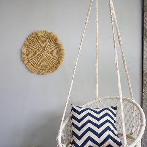 May include: A white macrame hanging chair with a blue and white chevron patterned pillow. The chair has a fringe border and is hanging from a white ceiling.