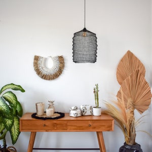 May include: A wooden table with two drawers and a black metal frame. On the table are two white ceramic candles, a small pineapple-shaped candle holder, and three decorative boxes with geometric patterns. A large, woven wall hanging with white accents is on the wall behind the table. A black metal pendant light hangs from the ceiling above the table.