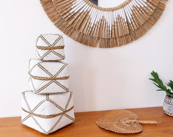 May include: Three white beaded boxes with gold geometric designs stacked on top of each other. The boxes are on a wooden surface with a woven fan and a potted plant in the background.