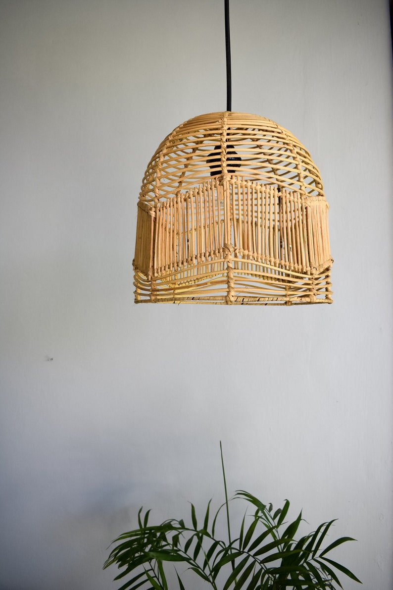 May include: A woven wicker pendant light fixture hangs from a black cord against a white wall. The light fixture is round and has a natural, light brown color.