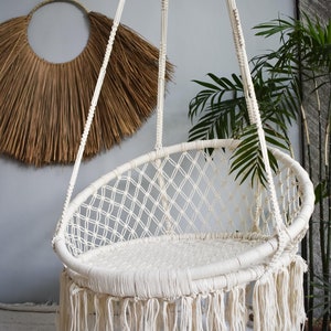 May include: A white macrame hanging chair with a fringe border. The chair is made of thick cotton rope and has a woven design.