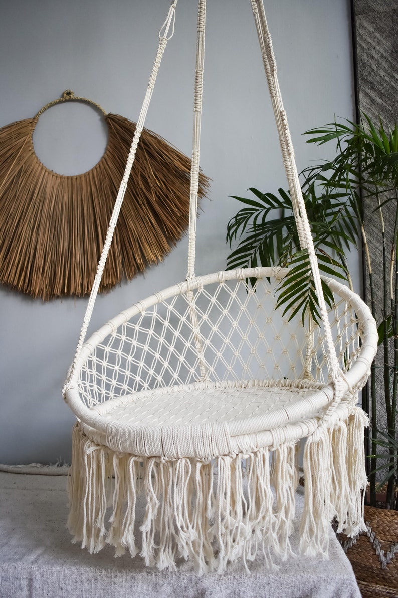 May include: A white macrame hanging chair with a fringe border. The chair is made of thick cotton rope and has a woven design.