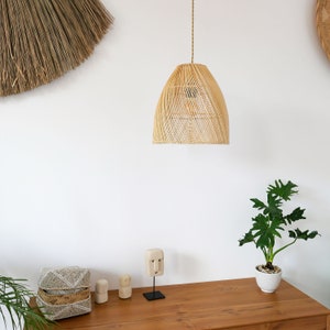 Pu&ograve; includere: Un lampadario a sospensione in rattan intrecciato &egrave; appeso a un soffitto bianco. Il lampadario &egrave; di colore beige naturale e ha un paralume rotondo a forma di cupola. Il lampadario &egrave; appeso sopra un tavolo di legno con una piccola pianta e oggetti decorativi.