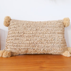 May include: A rectangular woven pillow with a natural straw-like texture and pom-poms on each corner. The pillow is a light beige color and is sitting on a wooden surface.