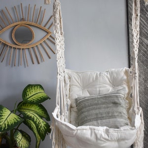 Puede incluir: Una silla colgante de macram&eacute; blanca con un coj&iacute;n blanco y una almohada a rayas grises. La silla est&aacute; colgada de una barra de madera y est&aacute; frente a una pared azul claro.