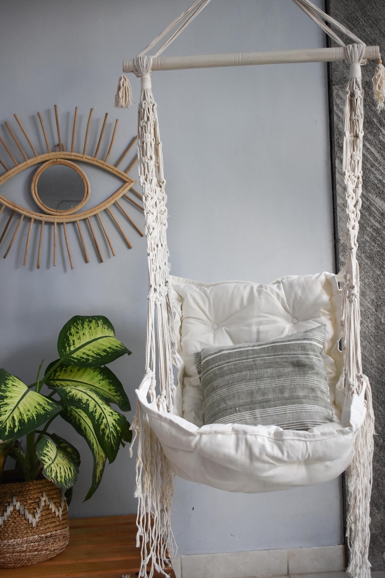 Puede incluir: Una silla colgante de macram&eacute; blanca con un coj&iacute;n blanco y una almohada a rayas grises. La silla est&aacute; colgada de una barra de madera y est&aacute; frente a una pared azul claro.