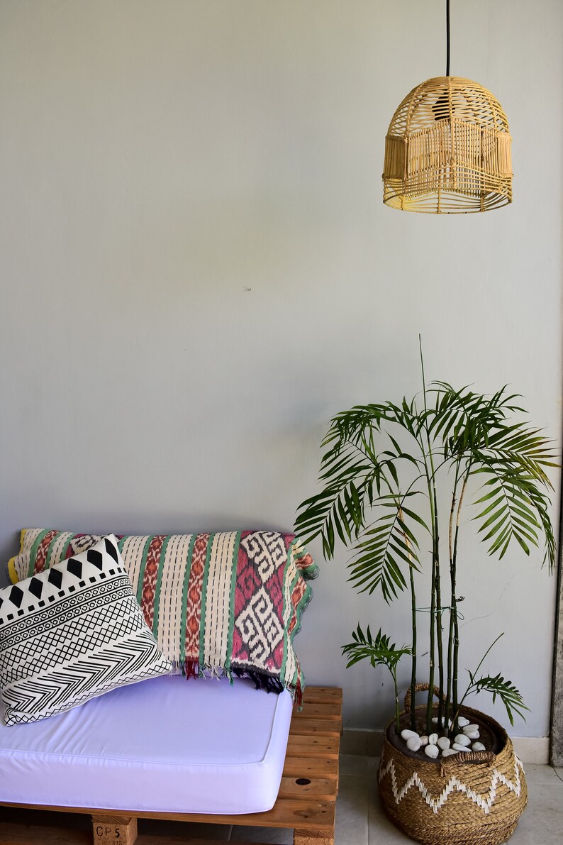May include: A white pallet couch with a purple cushion and two patterned pillows. A woven wicker lampshade hangs from the ceiling. A potted palm plant sits on the floor next to the couch.