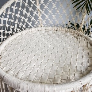 May include: A white macrame hanging chair with a woven seat and fringe detail. The chair is made of natural fibers and has a relaxed, bohemian style.