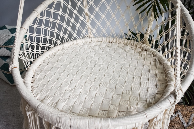 May include: A white macrame hanging chair with a woven seat and fringe detail. The chair is made of natural fibers and has a relaxed, bohemian style.