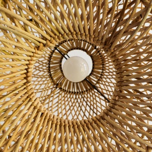 May include: A close-up view of a woven wicker lampshade with a white light bulb inside. The lampshade is made of natural materials and has a warm, rustic look.