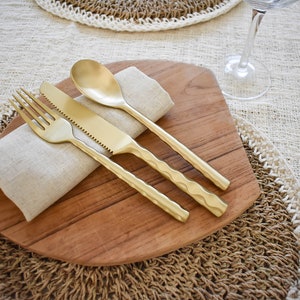 Puede incluir: Un juego de cubiertos de color dorado, que incluye un tenedor, un cuchillo y una cuchara, descansa sobre una tabla de servir de madera con una servilleta de lino. Los utensilios tienen un diseño texturizado y martillado. El conjunto está sobre un mantel individual tejido y un mantel texturizado.