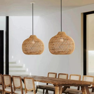May include: Two woven rattan pendant lights hang over a wooden dining table with six chairs. The chairs are made of woven rattan and have a natural wood frame.