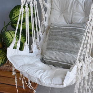 Puede incluir: Silla hamaca de macram&eacute; blanco con un coj&iacute;n blanco y una almohada a rayas grises. La silla est&aacute; colgada del techo y est&aacute; rodeada de plantas verdes.