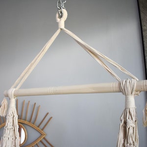 Puede incluir: Una hamaca de macram&eacute; blanca colgando de una barra de madera con borlas en los extremos. La barra est&aacute; unida a una cadena.