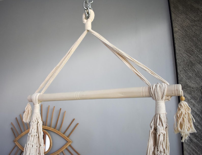 Puede incluir: Una hamaca de macram&eacute; blanca colgando de una barra de madera con borlas en los extremos. La barra est&aacute; unida a una cadena.