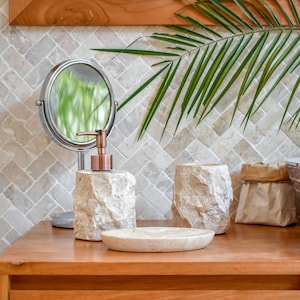 Puede incluir: Juego de accesorios de baño sobre una superficie de madera. El juego incluye un dispensador de jabón con bomba de cobre, un jabonera redonda y un vaso, todos hechos de piedra texturizada de color claro. Un pequeño espejo y una hoja de palma verde en el fondo.