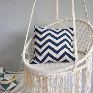 May include: A white macrame hanging chair with a blue and white chevron patterned pillow. The chair has a fringe around the bottom.